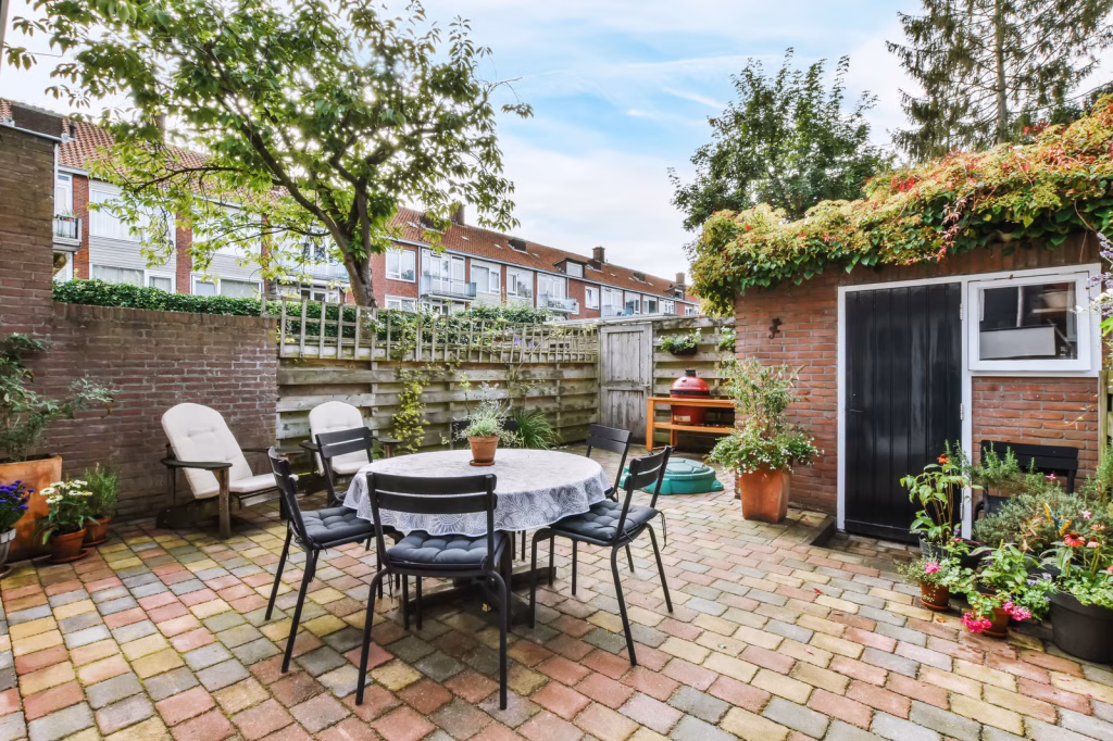 A multi-colored brick patio is set at the edge of a yard. The patio is edged by a brick wall, potted plants, a table with chairs, and a tablecloth in the middle–outdoor hardscaping ideas
